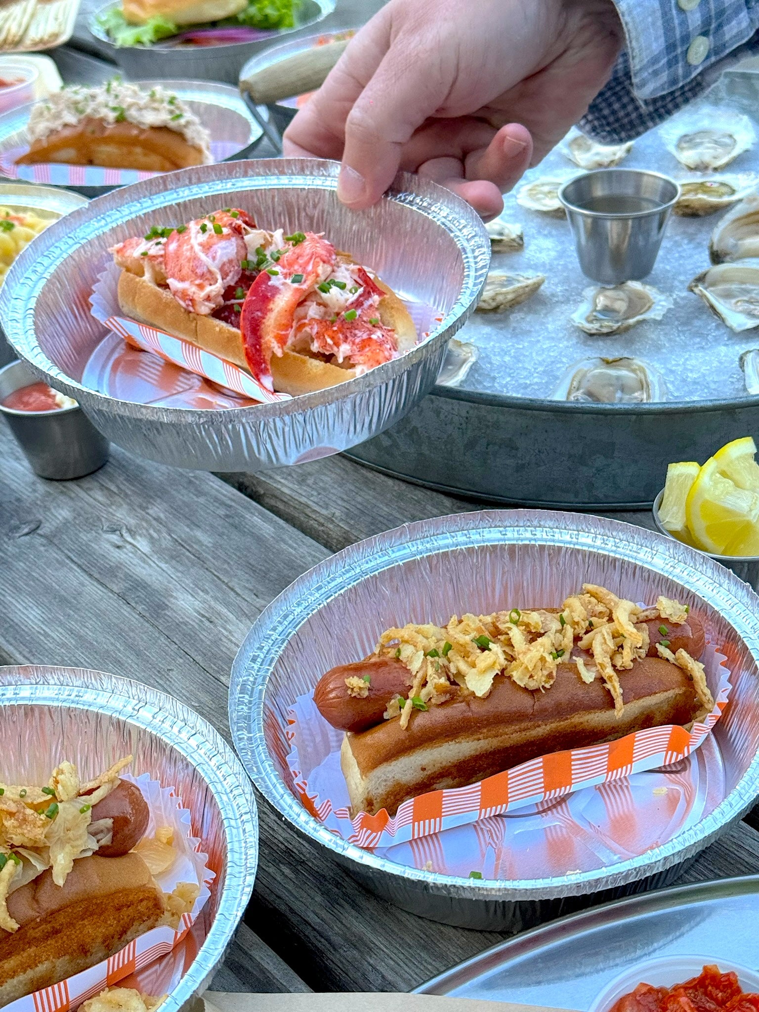 Hot dogs in foil containers on a wooden table with various condiments.