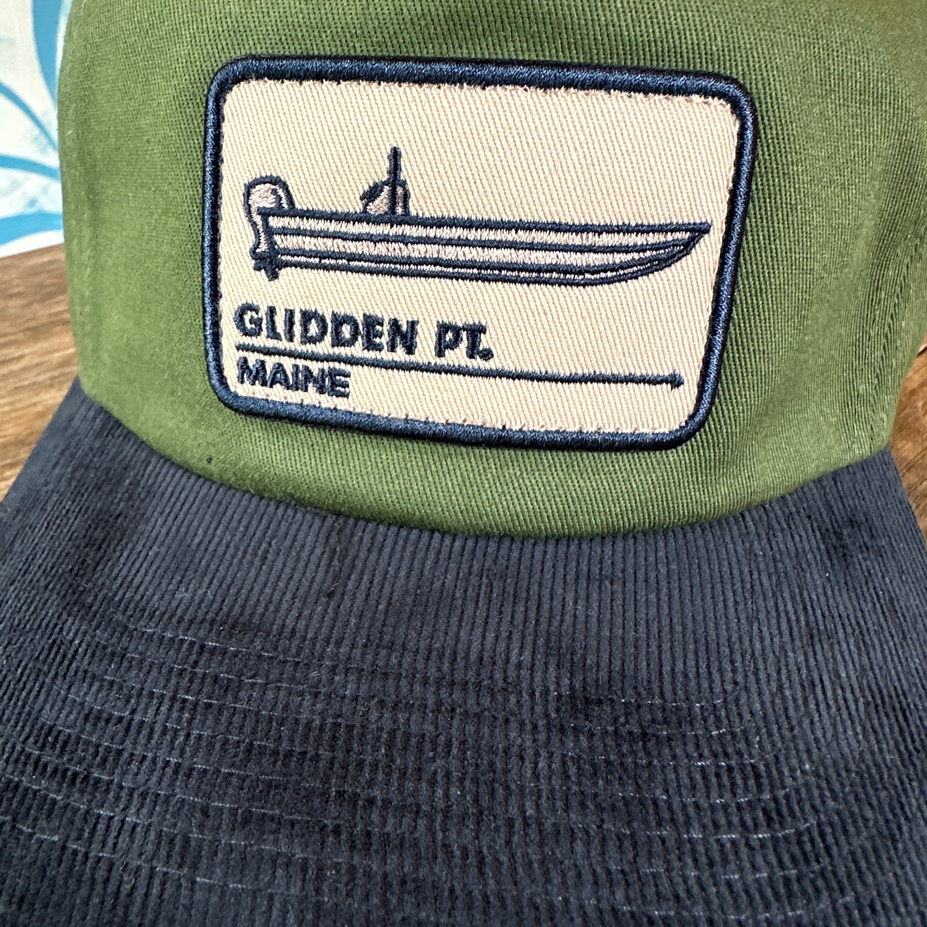 Green cap with a patch featuring 'Glidden Pt. Maine' on a wooden surface.