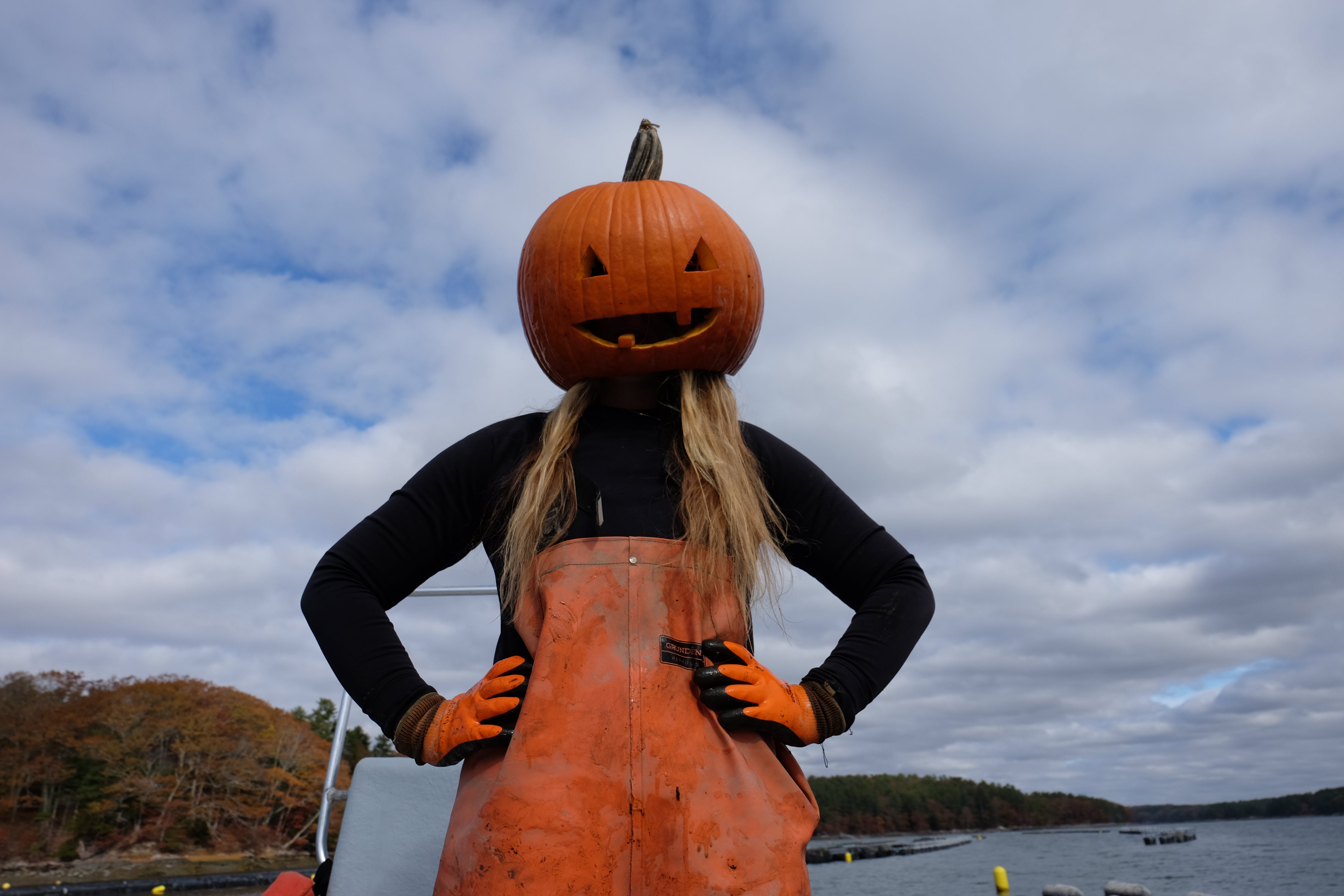 Person wearing a pumpkin head costume with a black shirt and orange apron, standing outdoors against a cloudy sky.