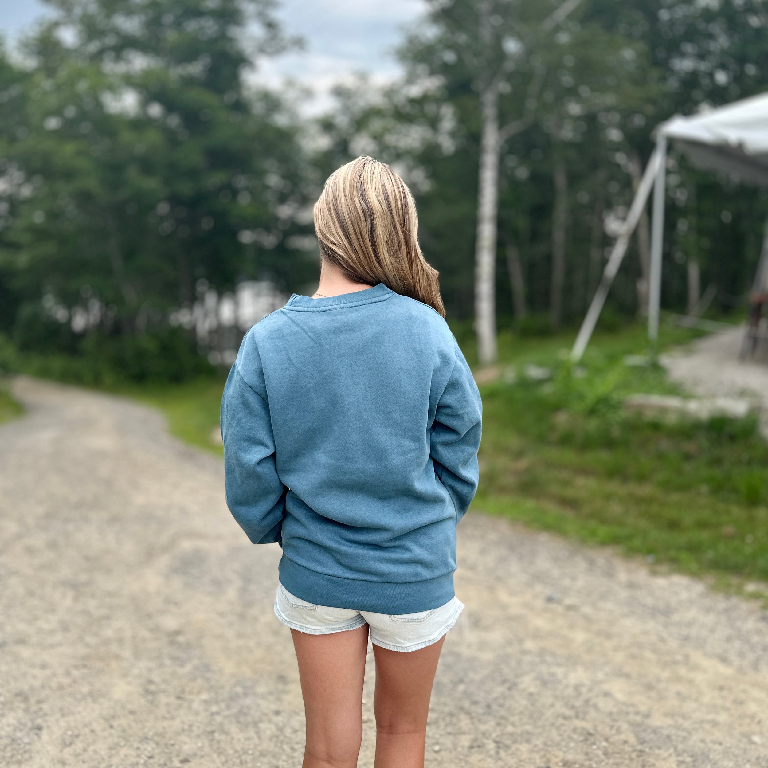 Person wearing a blue sweatshirt and white shorts standing on a gravel path with trees in the background