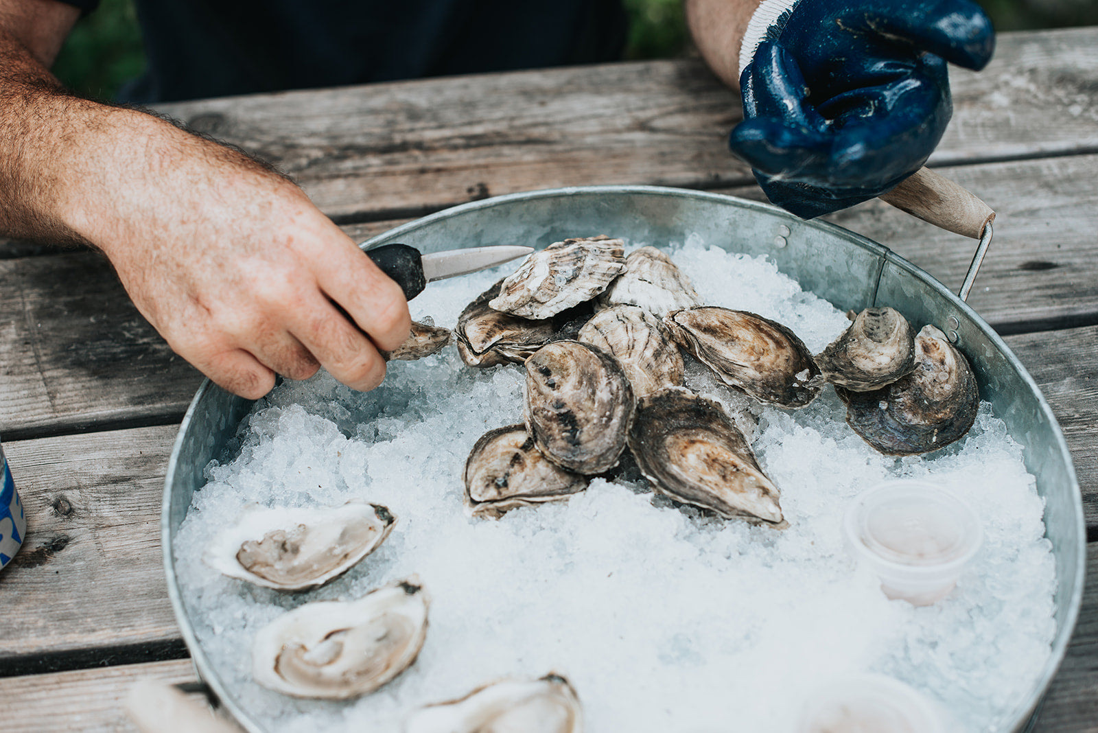 Person handling oysters on a bed of ice in a metal tray outdoors.
