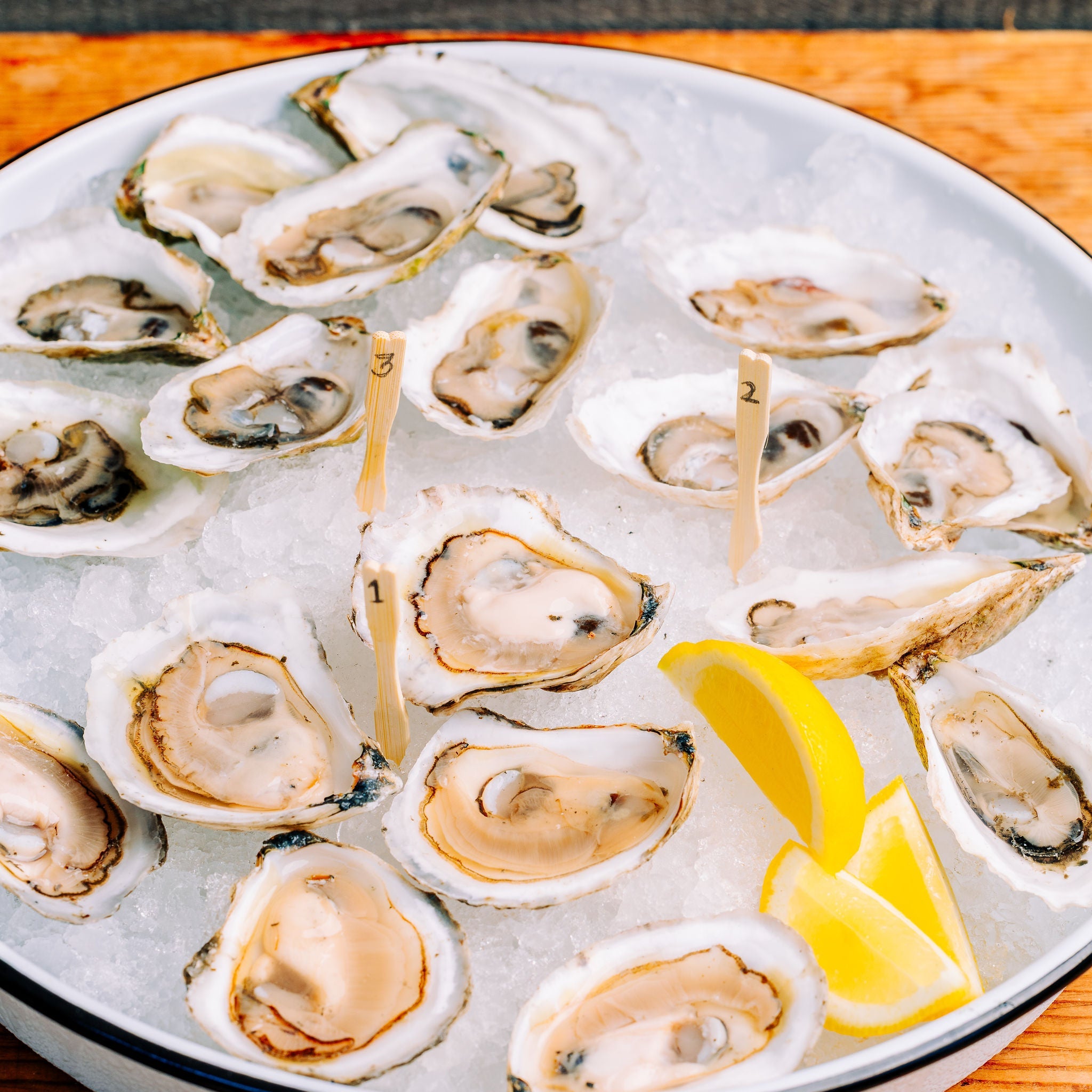 A white oval platter filled with raw oysters on ice, garnished with two lemon wedges, set on a wooden surface.
