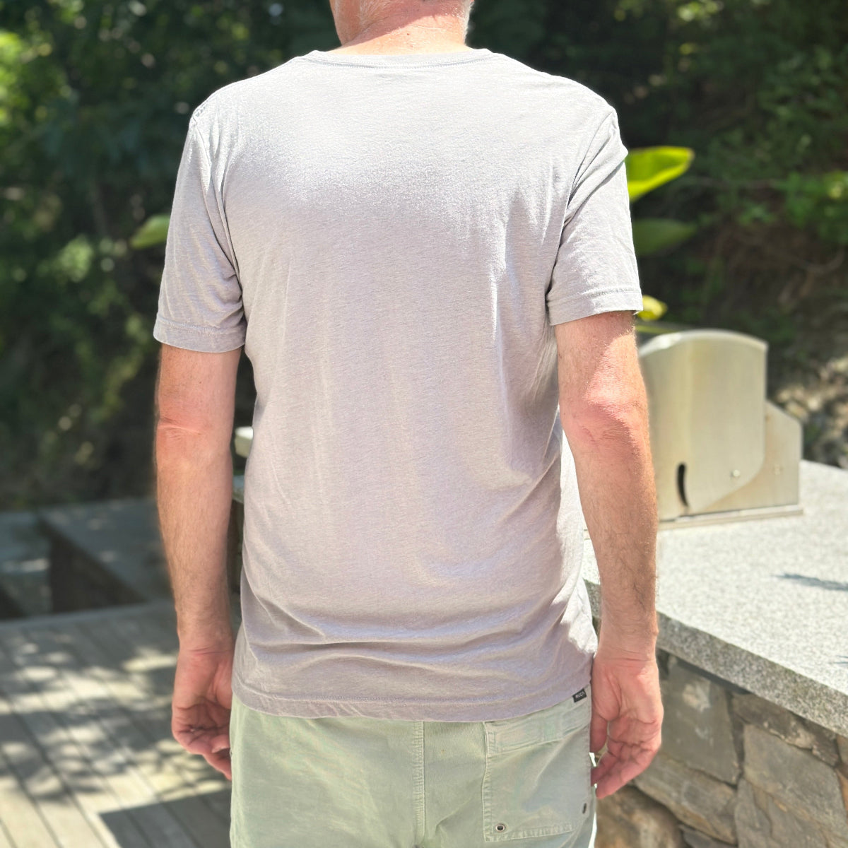 A man viewed from behind wearing a light beige short-sleeve crewneck t-shirt and light green pants, standing outdoors on a stone deck with green foliage in the background.