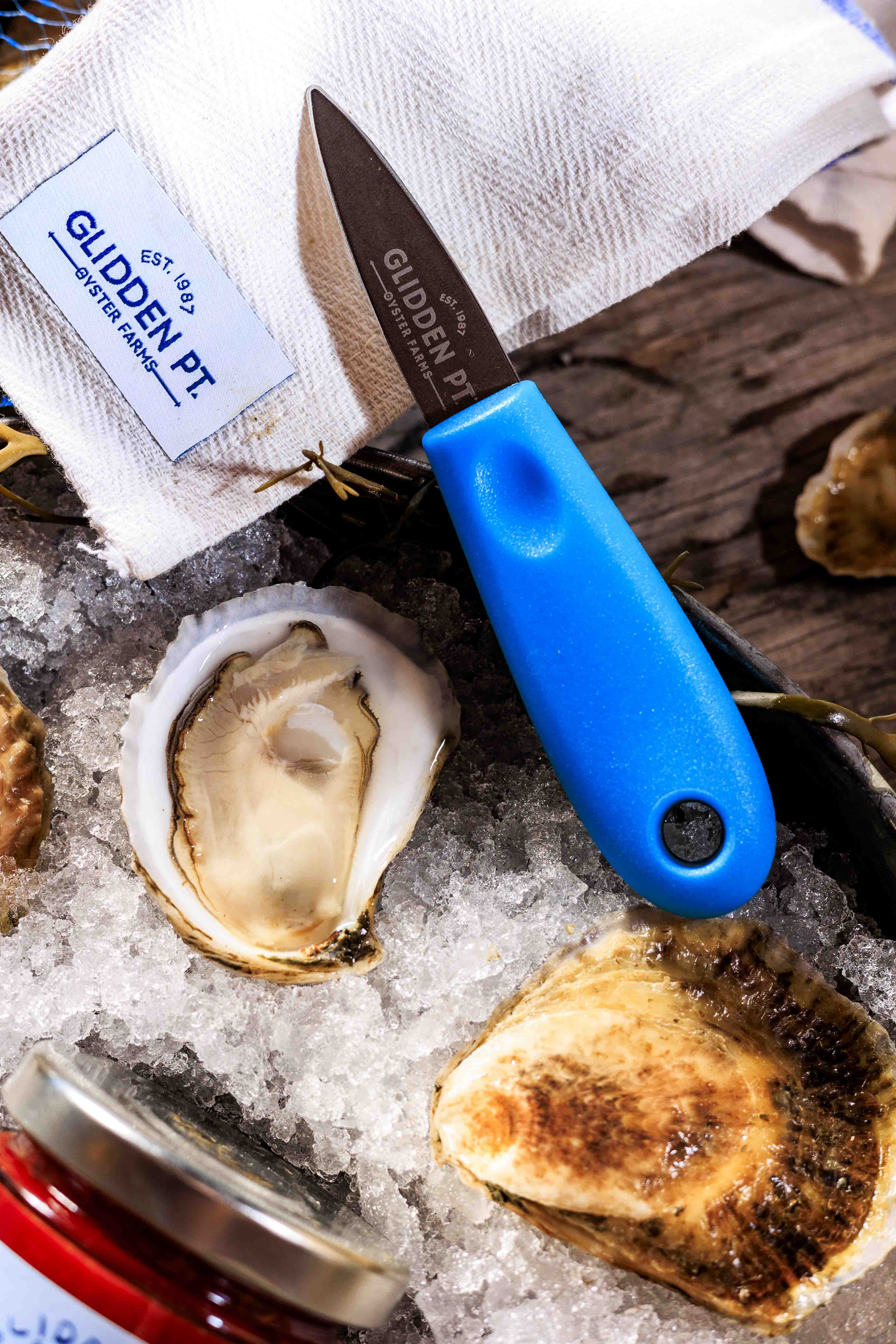 Oyster shucking knife with blue handle on ice next to oysters and a jar of mignonette.
