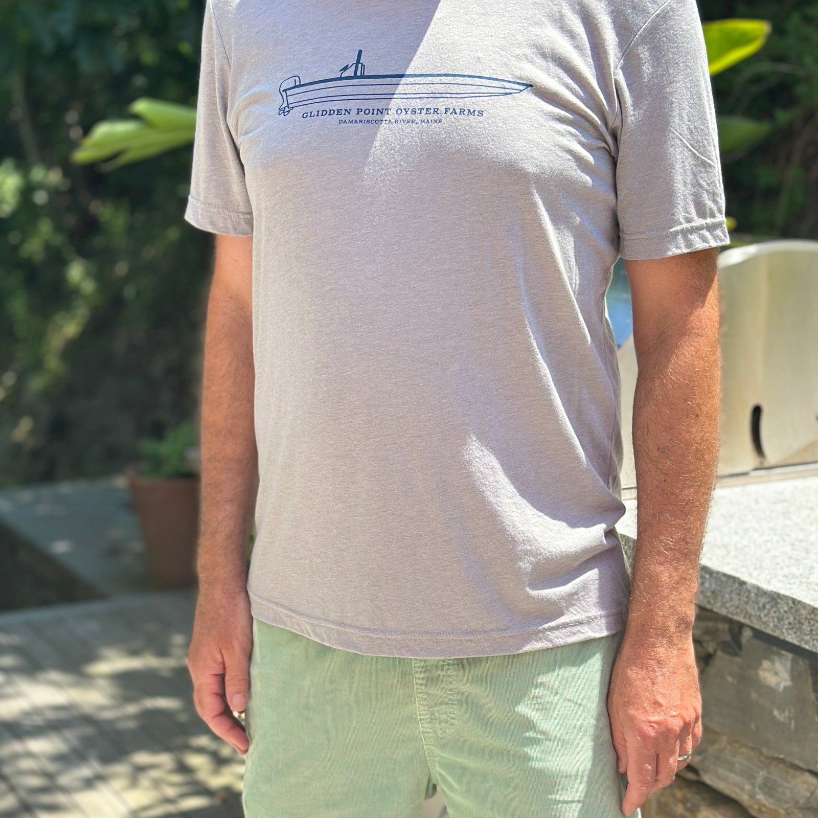 A man wearing a light gray heather crewneck t-shirt with a blue boat graphic and pale green shorts, standing on a wooden deck outdoors.