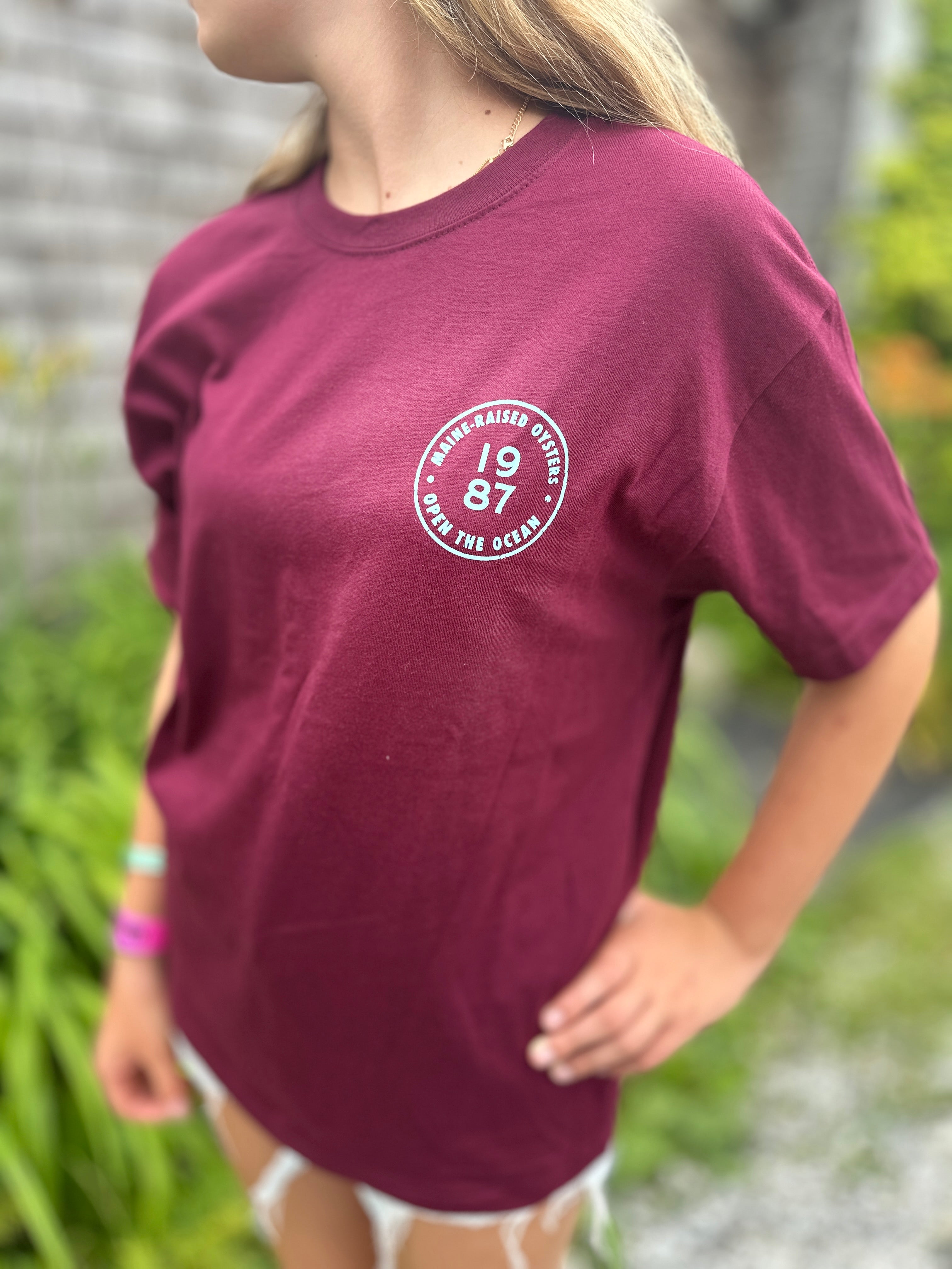 Person wearing a maroon t-shirt with a circular logo outdoors