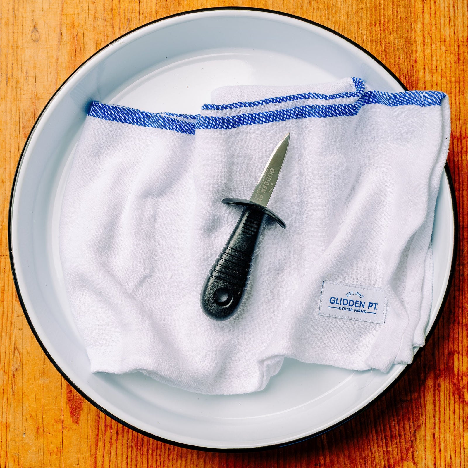 A white cloth with blue stripes and a black-handled knife resting on top, placed inside a white oval plate on a warm wooden surface. A small label on the cloth reads 'The Glidden Pt'.