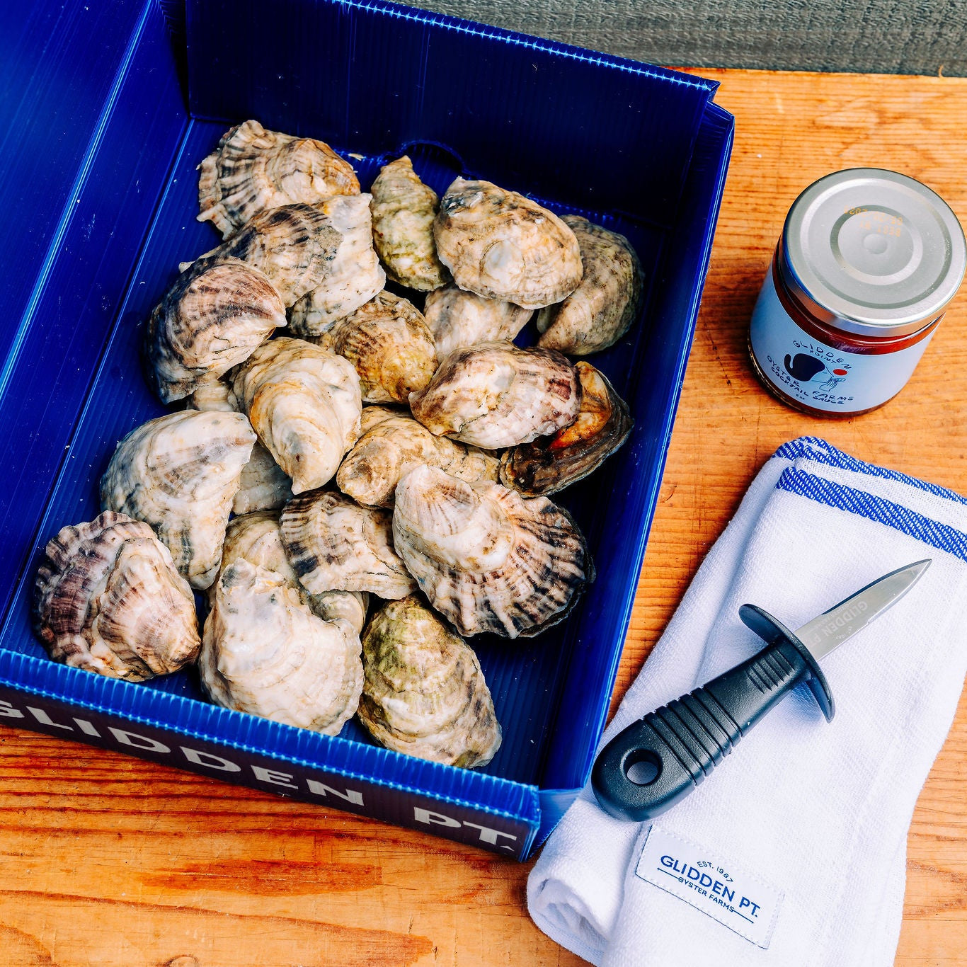 A blue cooler box filled with raw oysters on a wooden table, alongside a small can of sauce, a white napkin, and a black-handled knife.