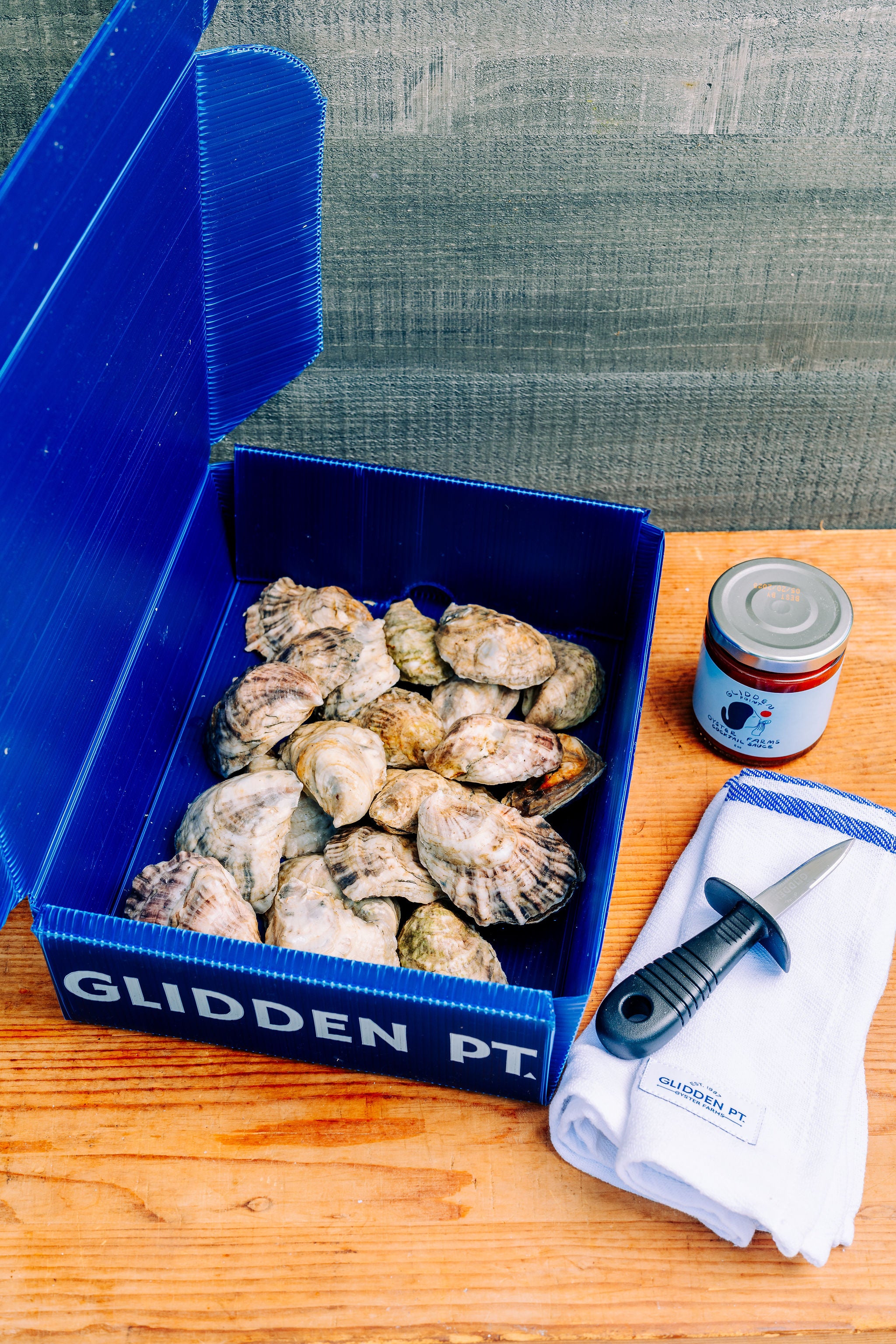 An open blue GLIDDEN PT. clam box filled with clams on a wooden table, alongside a small jar and a white towel with a knife.