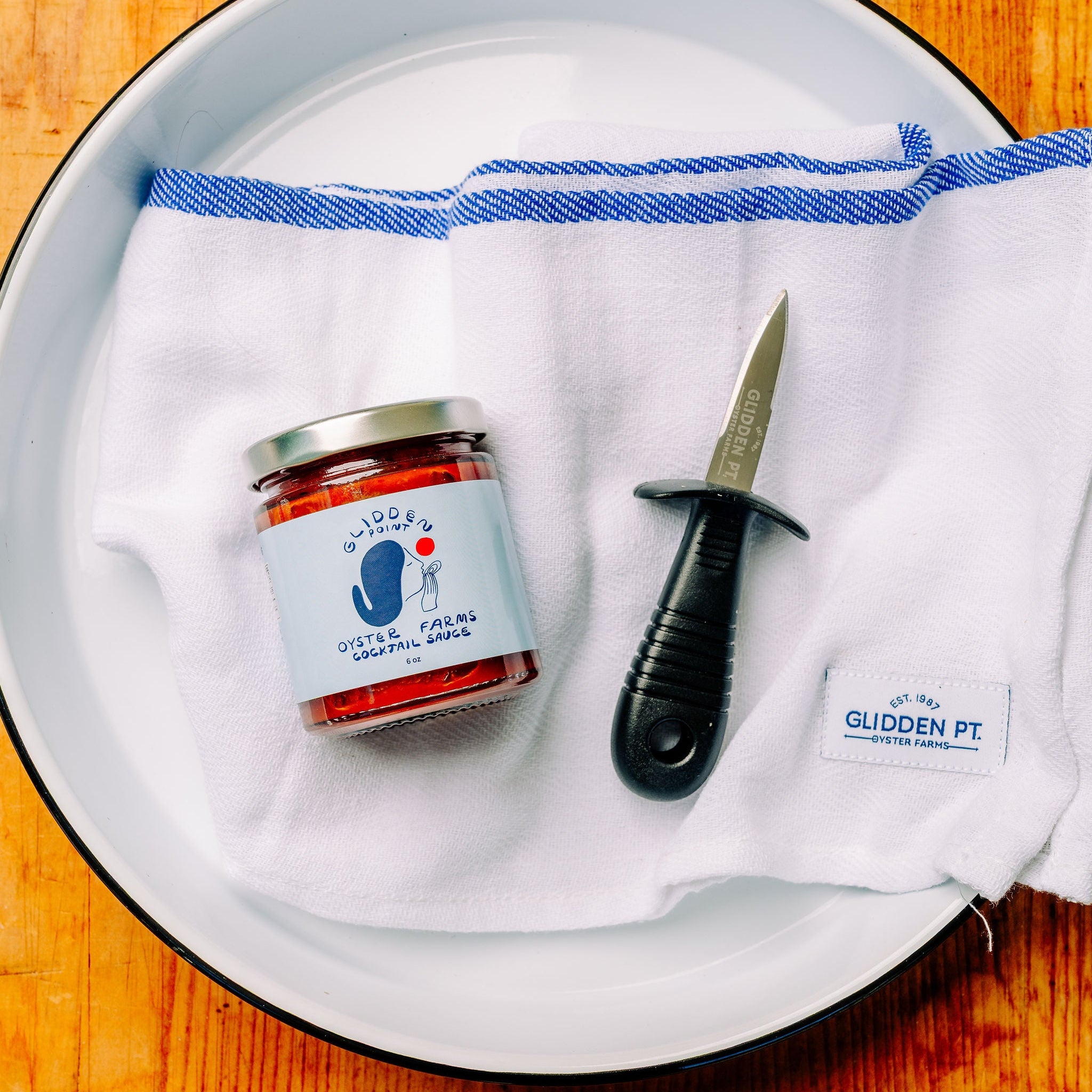 A small glass jar of red pepper sauce with a white label sits on a white cloth inside a white enamel plate, alongside a small knife and a white tag reading 'Glidden Point. The plate is placed on a warm wooden table.